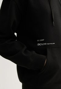 Felpa nera con cappuccio e tasca frontale, con testo bianco: "IDENIM TOM TAILOR" e "EST. 2007." Realizzata in cotone, con polsini a costine.