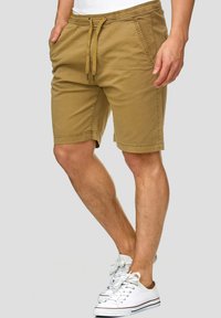 Shorts beige avec une taille élastique, cordon de serrage à l'avant et poches latérales. Portés avec des baskets blanches, mettant en avant un design décontracté et fonctionnel.