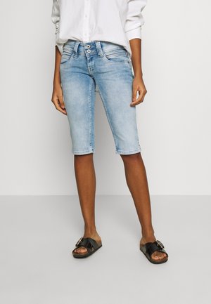 Persona con pantalones cortos vaqueros azul claro hasta la rodilla, camisa blanca de manga larga y sandalias negras sin talón con hebillas sobre un fondo liso.