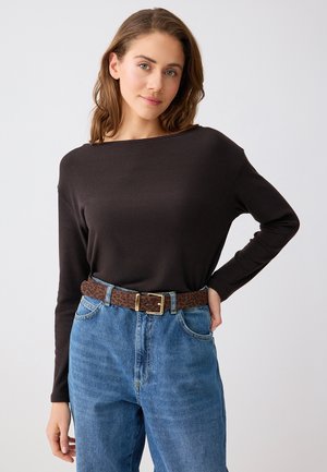 Jonge vrouw met lichtbruin haar, een zwart shirt met lange mouwen, high-waisted blauwe spijkerbroek en een bruine riem met luipaardprint.