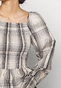 Ruudullinen yläosa, jossa on neliömäinen kaula-aukko, pitkät rypytetyt hihat ja teksturoitu elastinen smokkikangas. Värit ovat beige, musta ja harmaa erilaisissa kuvioissa.