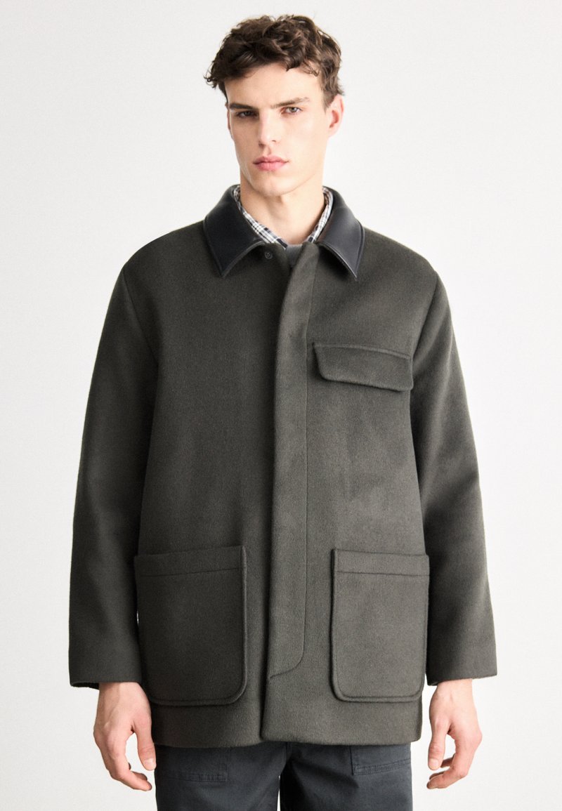 Παλτό σε ελιά πράσινο από μάλλινο μείγμα με γιακά από μαύρο δέρμα, διακοσμημένο με δύο εμπρόσθιες τσέπες και κρυφό μπροστινό κλείσιμο. Απλός και δομημένος σχεδιασμός.