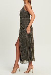 Kvinna klädd i en ärmlös, metallisk plisserad klänning med hög slits, i sällskap av beige högklackade sandaler, stående i profil.