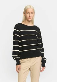 Zwarte en crèmekleurige gestreepte gebreide trui met een ronde halslijn en lange, iets opgeblazen mouwen; ribbelmanchetten en onderkant. Draagt over beige broeken.
