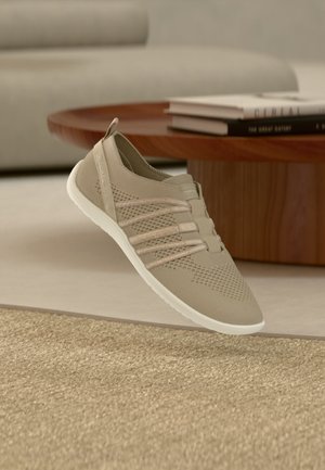 Šviesiai smėlio spalvos megztas slip-on stiliaus sportbačiai su elastinėmis juostelėmis ir balta pado dalimi, sklendžiantis virš tekstūruoto kilimo šalia medinio kavos staliuko su knygomis.