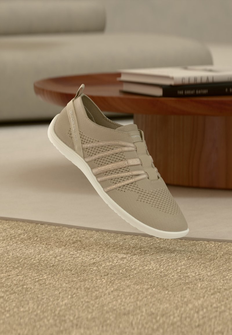 Ανοιχτό μπεζ πλεκτό παπούτσι τύπου slip-on με ελαστικές λωρίδες και λευκή σόλα που αιωρείται πάνω από υφασμένο χαλί δίπλα σε ξύλινο τραπεζάκι καφέ με βιβλία.