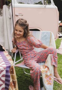 Bloemenpatronen jumpsuit in roze, blauw en oranje tinten; losse pasvorm met pofmouwen. Lichtroze slipper, zittend op een pastelstoel.