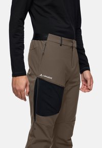 Bruine outdoorbroek met een zwarte zak, gemaakt van lichtgewicht materiaal. Kenmerkt zich door een tapered ontwerp en ritssluitingen aan de zijkant.