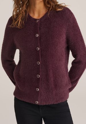 Femme portant un cardigan boutonné violet foncé, doux et duveteux, à manches longues, avec un pantalon noir sur fond uni.