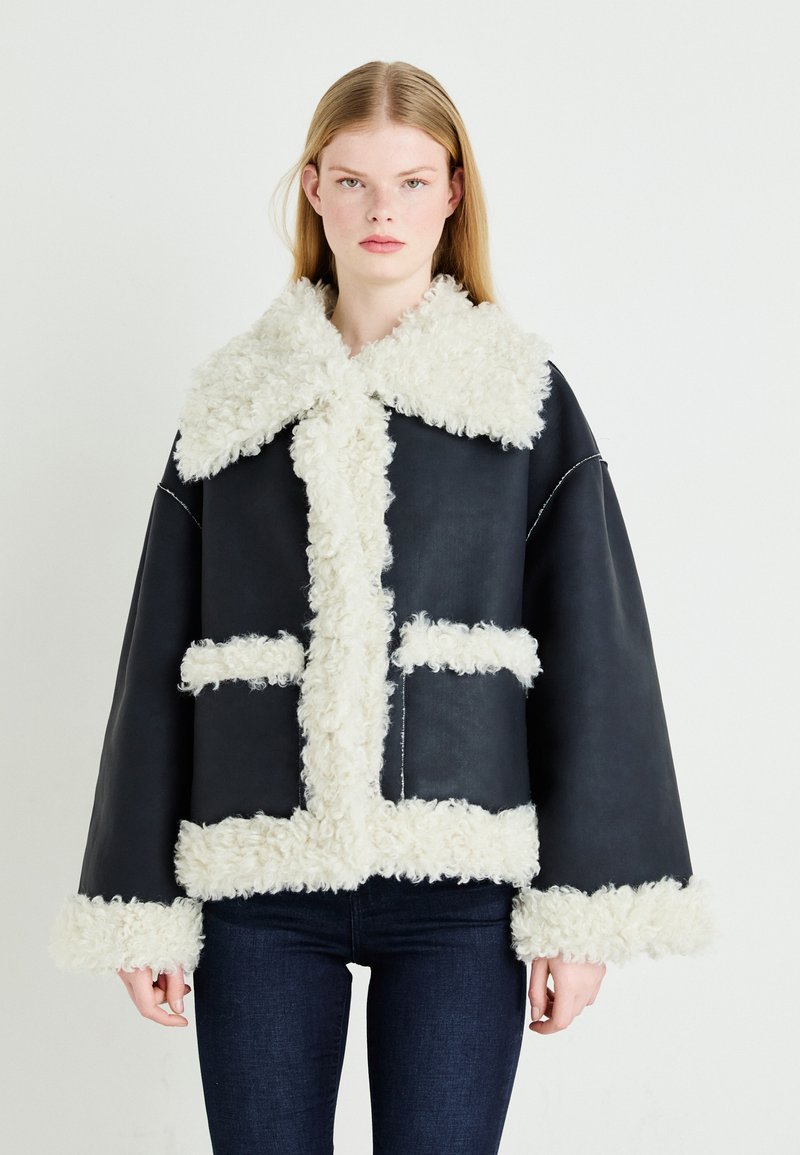 Veste en cuir noire avec col et bordure en fourrure blanche. Dotée de deux poches avant, d'une coupe carrée et d'une finition texturée.