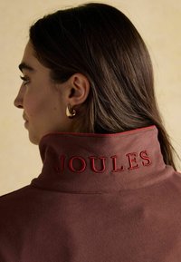 Hnedá bunda s vysokým golierom a červeným vyšívaním "JOULES". Mäkká textúra s kontrastným lemovaním goliera. Viditeľné zlaté kruhové náušnice.