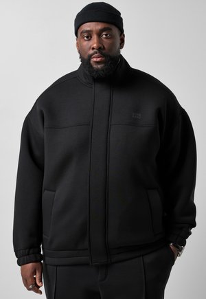 Mann mit Bart, der eine schwarze Mütze und eine schwarze Zip-up-Jacke trägt, mit den Händen teilweise in den Taschen, vor einem einfarbigen Hintergrund.