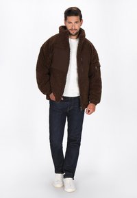 Mann in braunem Fleece-Jacke, weißem T-Shirt, dunkelblauer Jeans und weißen Sneakers steht vor einem weißen Hintergrund.