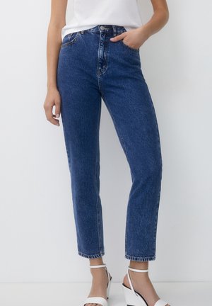 Femme portant un jean bleu taille haute avec poches, un top blanc sans manches et des sandales compensées blanches à bride à la cheville, debout contre un mur uni.