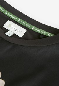 Černé tričko s hladkým povrchem. Má kulatý výstřih a zelenou etiketu s nápisem "by Ted Baker" a malým logem uvnitř límce.