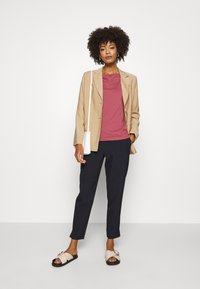 Beige bleiseri maroon t-paita ylleen, yhdistettynä tummiin housuihin ja vaaleisiin sandaaleihin, joissa on koristeellinen solki. Tausta on tasainen valkoinen.