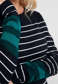 Námořnický pruhovaný svetr s bílými horizontálními pruhy a zeleně pruhovanými rukávy. Měkká pletenina s volným střihem.