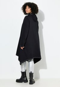 Zwarte oversized hoodie met langere achterkant, zilveren accentstiksels, gecombineerd met metallic zilveren leggings en zwarte enkellaarsjes.
