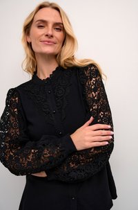 Svart blus med intrikata spetslånga ärmar, knappad framtill, dekorativ spetsdetalj vid kragen, och åtsittande manschetter.