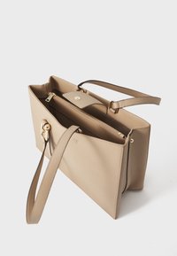 Sac fourre-tout beige, fabriqué en matériau lisse. Équipé de quincaillerie dorée, d'un compartiment zippé et de doubles poignées pour le transport.