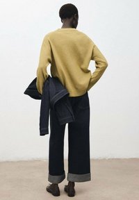 Maglione giallo senape, pantaloni in denim scuro a gamba larga con risvolti, tiene in mano una giacca di denim. Tessuto liscio e design semplice.