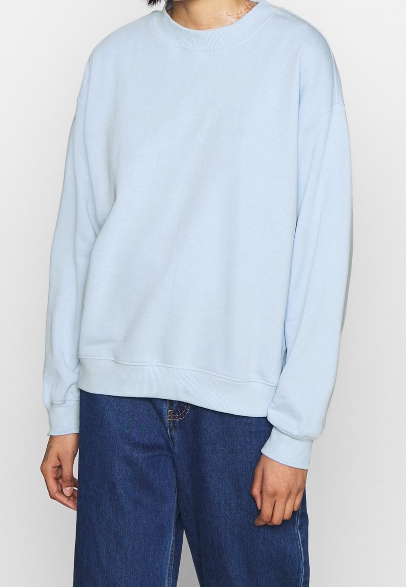 Person iført en simpel lyseblå sweatshirt med lange ærmer, kombineret med mørkeblå denimjeans, stående mod en hvid baggrund.