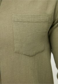 Camisa verde oliva de tela texturizada, con un bolsillo en el pecho y un cierre frontal con botones. El primer plano muestra los detalles del tejido y la costura.