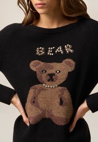 Suéter negro con un diseño de osito de peluche marrón texturizado y acentos de pedrería. La palabra "OSO" está adornada con joyas por encima del oso.