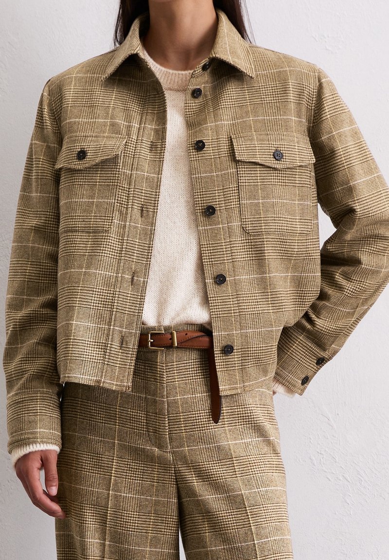 Femme portant une veste à carreaux beige avec des boutons noirs, un pantalon à carreaux assorti, une ceinture marron et un pull en laine crème sur fond blanc.