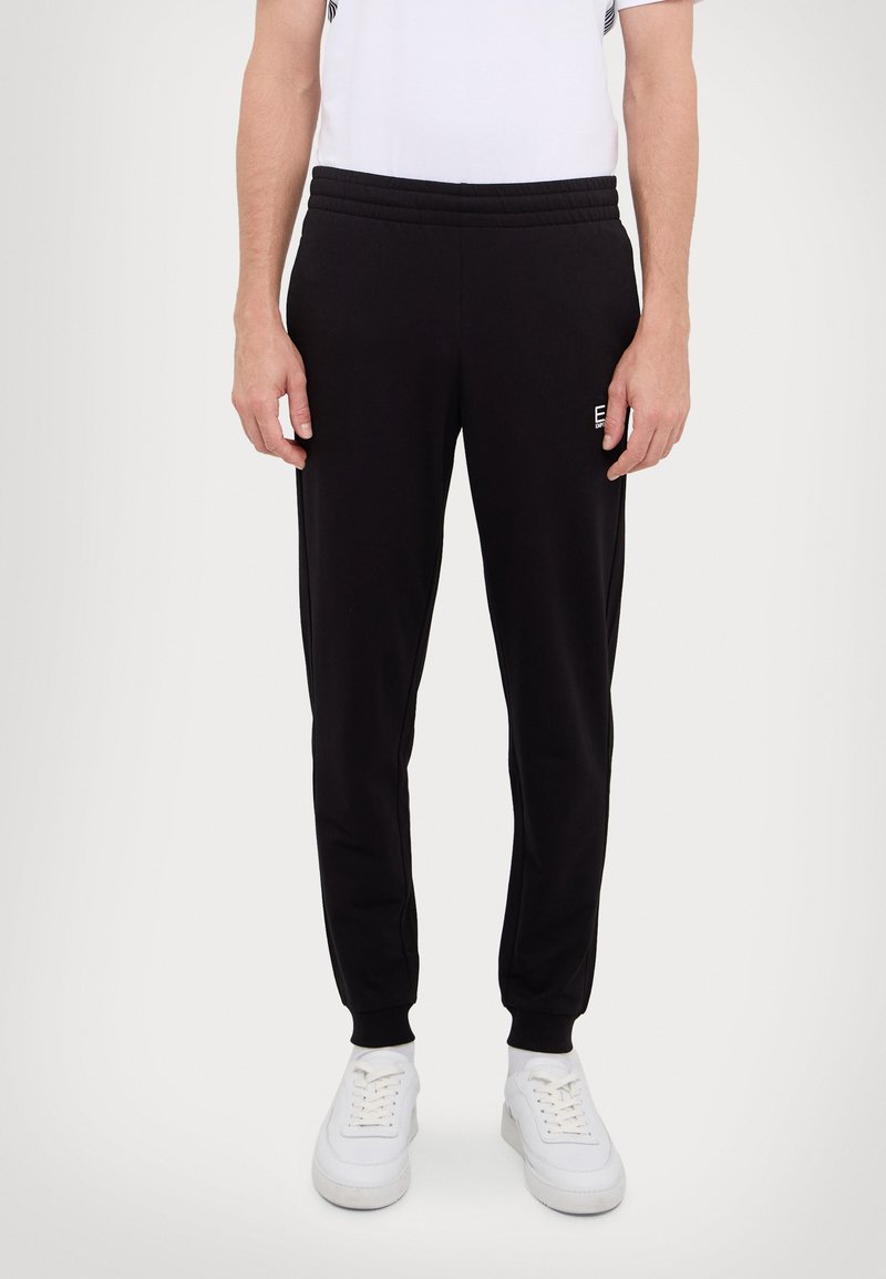 Zwarte sweatpants met een elastische tailleband en boorden. Bevat een klein logo op de dij. Draagt met witte sneakers. Minimalistisch ontwerp.