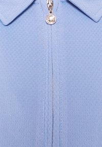 Ljust blå polo skjorta med en texturerad tyg, med halvdragkedja och rundad krage. Silverfärgad dragkedjehalare med en cirkulär detalj.