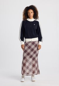 Marineblauen Reißverschluss-Pullover mit weißen Streifen und gerippten Bündchen, kombiniert mit einem langen Karorock in Rot und Creme sowie weißen Turnschuhen.
