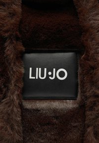 Brun syntetisk pels med en glat sort læderlap, der viser "LIU·JO" i hvide bogstaver. Teksturen ser blød og tæt ud.