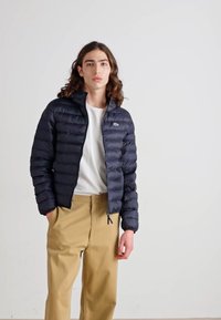 Mörkblå puffjacka med en quiltad design, lätt material, hög krage och framficka, bärs över en vit skjorta och beige byxor.