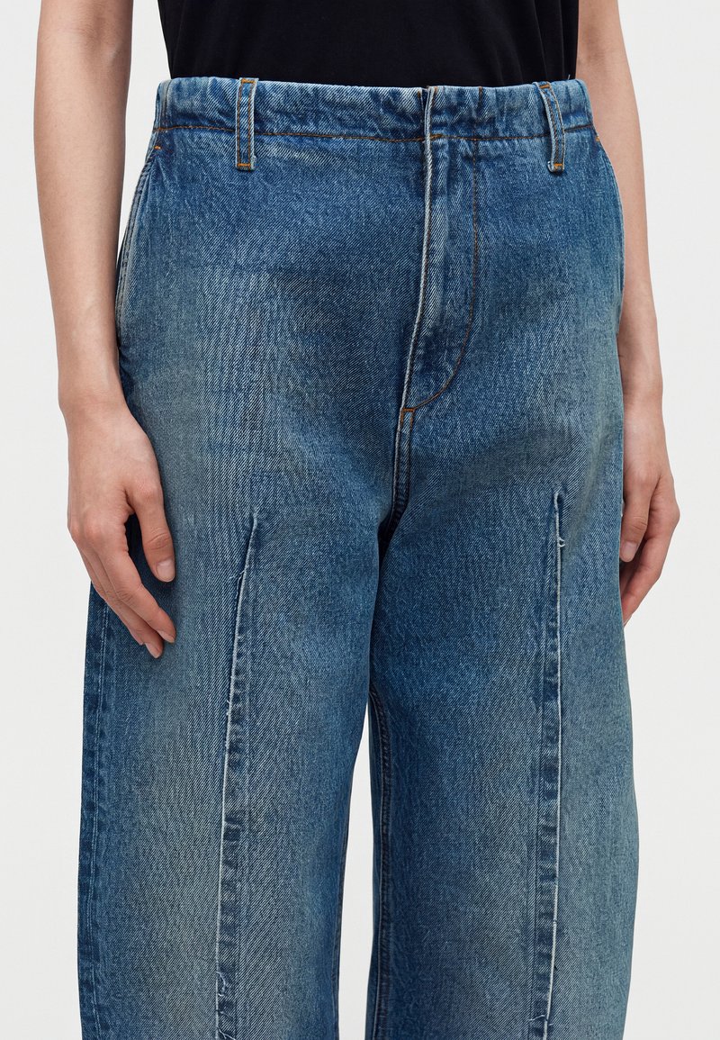 Person trägt locker sitzende blaue Jeans mit vorderer Naht und ein schwarzes, eingestecktes Shirt, steht vor einem schlichten, hellen Hintergrund.