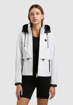 Ung kvinde iført en hvid hættetrøje med lynlås over en sort tætsiddende top og sorte cykelshorts, stående mod en ensfarvet baggrund.