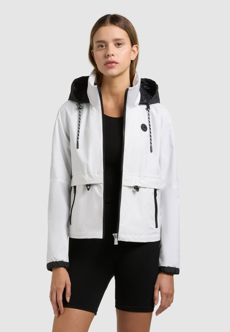 Jeune femme portant une veste blanche à capuche zippée par-dessus un haut noir ajusté et un short de cyclisme noir, debout devant un fond uni.