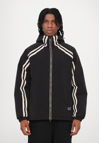 Črna jakna s kapuco, z belimi in zlato črtastimi poudarki. Narejena iz mehke tkanine z zadrgo in stranskimi žepi.