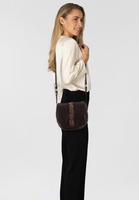Dunkelbraune Leder-Crossbody-Tasche mit dekorativen Perlen entlang der Mitte, geschwungener Form und langem verstellbarem Tragegurt.