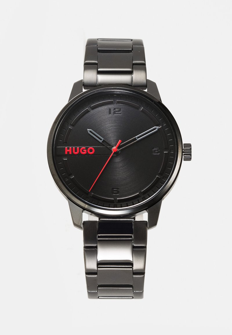 Relógio de pulso preto com bracelete em aço inoxidável, mostrador redondo, design minimalista, detalhes vermelhos para o logótipo e o ponteiro dos segundos, textura escovada.