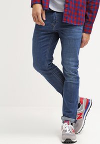 Blå denimjeans med slim fit, med subtila slitningar och sydda detaljer. I kombination med grå och röda träningsskor.