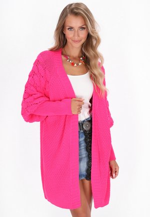 Vrouw in een felroze dikke gebreide cardigan over een witte top en een spijkerrok met een zwarte riem en een kleurrijke kralenketting.