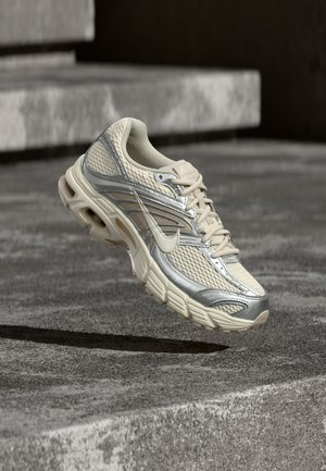 Beige and silver Nike running shoe with mesh and lace details hovering above concrete steps.