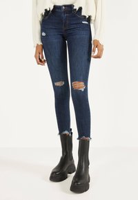 Jeans de tiro bajo ajustados de color azul oscuro con roturas y dobladillos deshilachados, combinados con botas negras de tobillo gruesas y un suéter texturizado color crema.