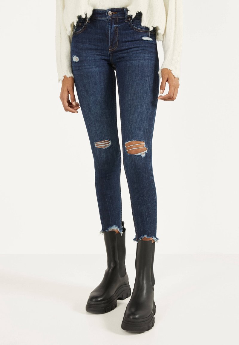 Jeans de tiro bajo ajustados de color azul oscuro con roturas y dobladillos deshilachados, combinados con botas negras de tobillo gruesas y un suéter texturizado color crema.