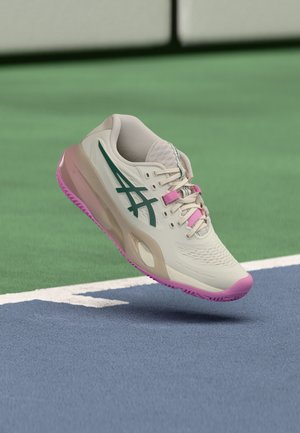 Witte en roze sportschoen met groen logo die boven een tennisbaan zweeft vlak bij de achterlijn.