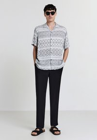 Chemise à manches courtes à motifs noir et blanc, pantalon noir et sandales noires avec détails en nœud, portées avec des lunettes de soleil.
