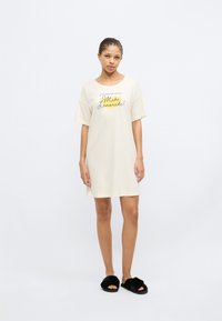 Φόρεμα από βαμβακερό ύφασμα εκρού χρώματος με άνετη εφαρμογή, κοντά μανίκια και τυπωμένο σύνθημα. Μαύρα πλαστικά σανδάλια τύπου σλίπ. Απλός και καθημερινός σχεδιασμός.