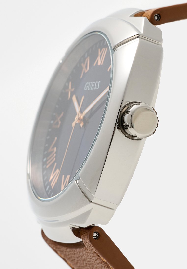 Reloj Guess con caja de metal plateado, esfera azul oscuro, marcadores de números romanos y correa de cuero marrón texturizado. Cuenta con una corona en el lateral.