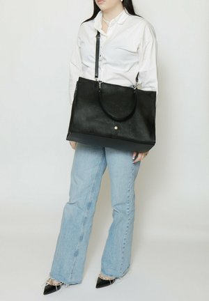 Donna con camicia bianca e jeans azzurri chiari tiene una grande borsa tote nera in pelle con manici per la spalla e per la mano.